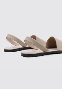 Sandalias deslizantes beige con diseño minimalista, parte superior de cuero suave y suela de goma negra texturizada. Presentan dos correas anchas para mayor estabilidad.