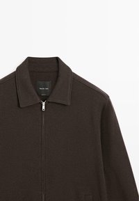 Ciemnobrązowa kurtka zapinana na zamek, wykonana z miękkiego materiału, z kołnierzem, szwami na ramionach i metką w środku. Prosty i minimalistyczny design.