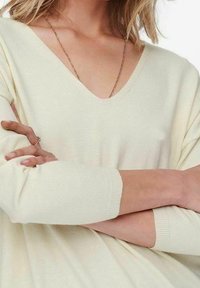 Femme portant un pull crème à col en V avec des manches longues, les bras croisés, et une délicate chaîne en or visible au niveau du col.