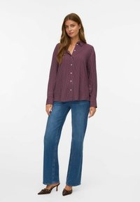 Kvinna klädd i en vinröd randig skjorta med knappar, blå vida jeans och bruna spetsiga klackskor, som står framför en enkel vit bakgrund.