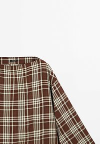 Chemise à manches longues à carreaux marron et blanc avec un col large, présentée sur un fond blanc uni.