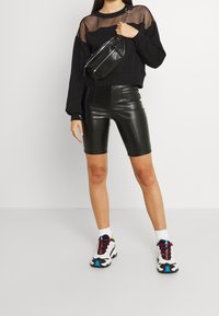 Sudadera negra recortada con superposición de malla, combinada con pantalones cortos de cuero sintético negros de cintura alta. El calzado presenta acentos coloridos y un diseño robusto.