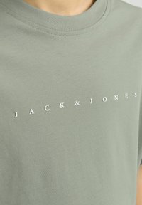 Olīvu zaļš t-krekls, kas izgatavots no kokvilnas, ar apaļu dekoltē. Priekšpusē centrā ir balts uzraksts ar zīmola nosaukumu "JACK & JONES".