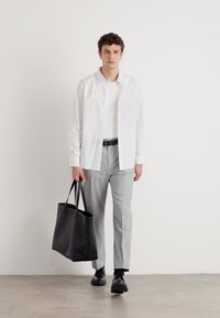 Weißes Hemd über einem weißen T-Shirt, graue Hose und schwarze Schuhe, hält eine schwarze Tote-Tasche. Minimalistisches Design und neutrale Farben.