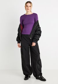 Μωβ μπλούζα με μακριά μανίκια, μαύρο μπουφάν τύπου bomber με γυαλιστερή υφή, μαύρο παντελόνι cargo με πολλαπλές τσέπες και μαύρα παπούτσια.