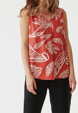 Kobieta ubrana w czerwoną bluzkę bez rękawów z abstrakcyjnymi białymi wzorami liści oraz czarne spodnie, stojąca na tle jednolitego jasnego tła.