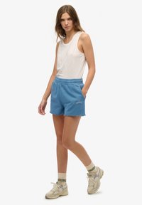 Jasnoszary top na ramiączkach, niebieskie krótkie spodenki dresowe z postrzępionym wykończeniem i przednimi kieszeniami, z haftowanym logo. Beżowe sneakersy z zielonym akcentem.