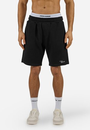 Man draagt zwarte geribde knielange shorts, witte merk-sokken, beige sneakers en witte onderbroek met tailleband, staand tegen een effen achtergrond.