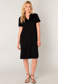 Zwarte knie-lange jurk met een V-hals en korte mouwen, gemaakt van zachte stof. Draagt zwarte sandaalhakken met bandjes, een gladde textuur en zonder patronen.