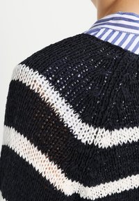 Cárdigan de punto con un patrón a rayas en azul marino, blanco y marrón. Textura de tejido abierto con un ajuste relajado y holgado.