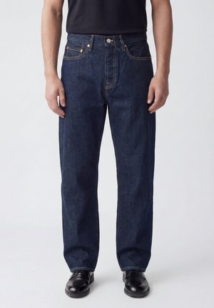 Uomo che indossa jeans blu scuro a gamba dritta, camicia nera e scarpe eleganti in pelle nera, in piedi davanti a uno sfondo chiaro uniforme.