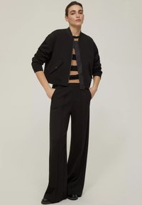 Giacca bomber nera corta con chiusura a zip, maniche lunghe e tasca laterale, abbinata a pantaloni neri a gamba larga e una maglietta a righe. Texture morbide.