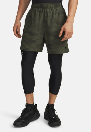 Hombre con pantalones cortos de camuflaje verde sobre mallas negras de compresión y zapatillas deportivas negras, de pie con los brazos relajados a los lados.