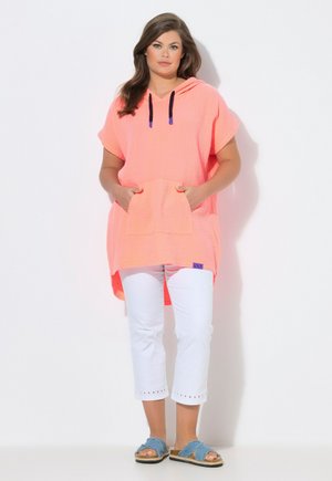 Frau trägt eine korallenfarbene Kapuzen-Tunika mit schwarzen Kordeln, weiße kurze Hosen und blaue Schlupfsandalen, sie steht vor einem weißen Hintergrund.