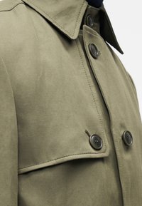 Palton verde măsliniu din material textil, cu guler, două nasturi negri și un buzunar lateral cu clapetă. Suprafață netedă, texturată.