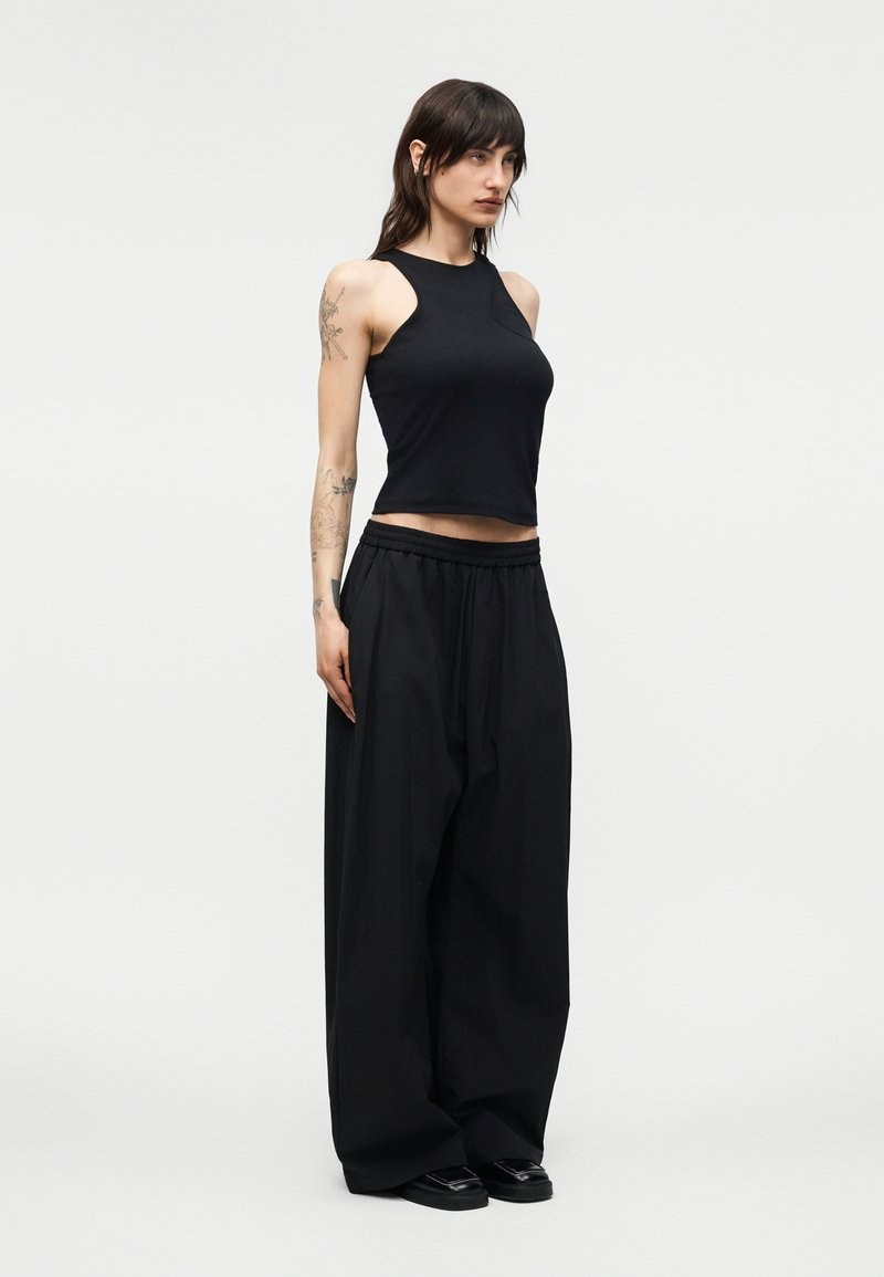 Femme aux cheveux foncés et tatouée, portant un crop top noir sans manches et un pantalon noir à jambes larges, debout devant un fond clair uni.