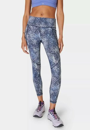 Vrouw met een high-waisted blauw-wit gespikkelde legging, een blauw crop top en grijze en paarse sportschoenen, staand tegen een witte achtergrond.