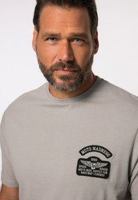 Šedé bavlněné tričko s kulatým výstřihem a černou vyšívanou nášivkou s textem, zobrazující křídlatý design na hrudi.