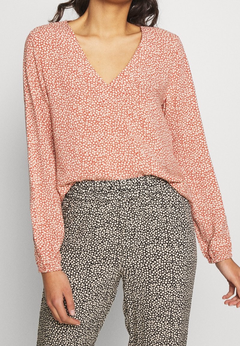 Femme portant un chemisier rose à manches longues avec col en V et un pantalon taille haute noir et blanc à motifs.