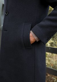 Cappotto di lana navy con finitura testurizzata, dotato di tasca frontale e polsini con bottone, mostrato con una mano appoggiata nella tasca.