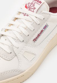 Biały skórzany sneaker z teksturowanymi akcentami, różową podszewką i czerwonym logo. Charakteryzuje się płaskimi sznurowadłami oraz perforowanymi detalami.