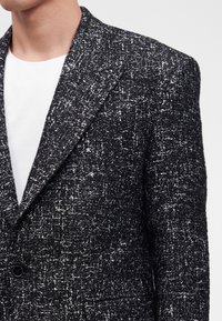 Gros plan d'une personne portant un blazer texturé noir et blanc par-dessus un t-shirt blanc uni à col rond.