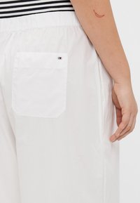 Hvite stoffshorts med elastisk midjebånd, en baklomme og et lite logo med røde, hvite og blå detaljer.