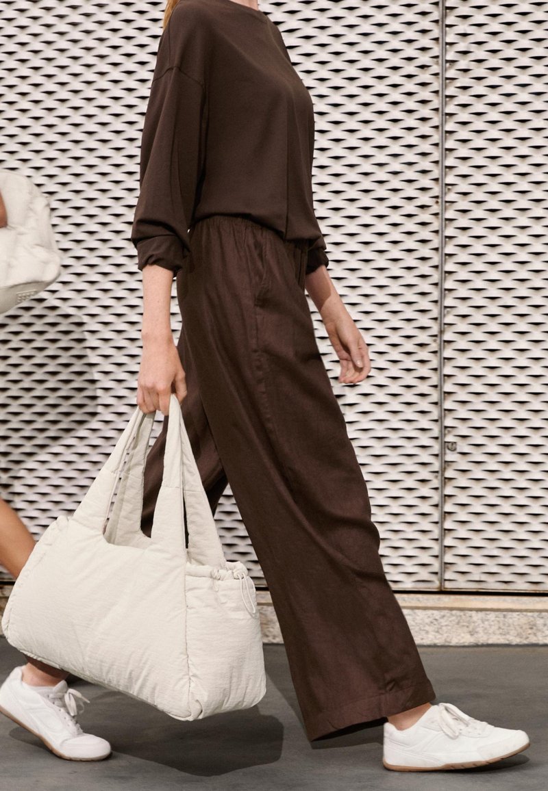 Brązowa, luźna bluzka i pasujące szerokie spodnie wykonane z miękkiego materiału. Biała pikowana torba na ramię i białe sneakersy z gumowymi podeszwami.