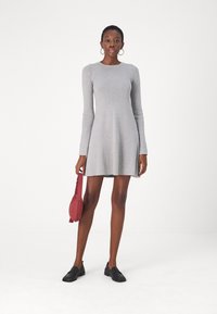 Grå randig långärmad klänning med utsvängd kjol. Modellen håller en röd handväska och bär svarta slippy-skor. Minimalistisk design och textur.