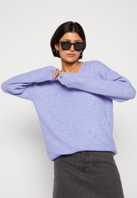 Pull en maille côtelée lavande clair avec un col en V, épaules tombantes et manches longues, associé à une jupe gris foncé. Des lunettes de soleil noires sont portées.