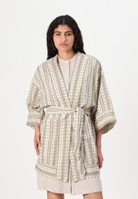 Prúžkovaný kimono vo svetlej smotanej farbe s čiernymi bodkami a červenými akcentmi; má široké rukávy, opasok a dĺžku do polovice stehna. Textúrovaná tkanina.