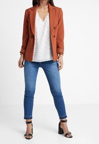 Rostfärgad dubbelknäppt blazer, vit topp med polka prickar, blå smala jeans och svarta klacksandaler med silverdetaljer.