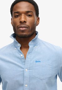 Lichtblauwe button-up blouse met een klein borstzakje, contrasterende witte knopen en een logo patch. Zachte stof met een gladde structuur.