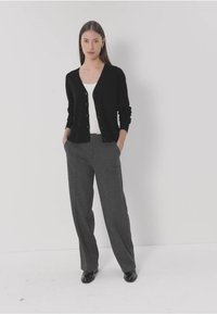 Zwarte cardigan met knopen, witte top eronder, en grijze wijduitlopende broek. Het model staat met de handen in de zakken voor een effen achtergrond.