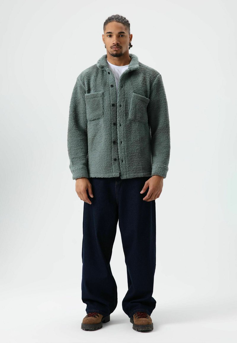 Groen, getextureerd fleecejack met een kraag, twee borstzakken en een voorzijde met knopen, gecombineerd met losse donkere spijkerbroeken en bruine schoenen.