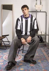 Jerseu supradimensionat cu dungi alb-negru, guler violet, pantaloni din denim gri, pantofi negri și accesorii din lanț argintiu, stând pe un scaun.