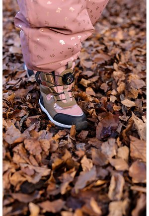 Bottes d'hiver roses et beige avec une semelle en caoutchouc noire et un laçage à cadran réglable, posées sur un lit de feuilles d'automne brunes.