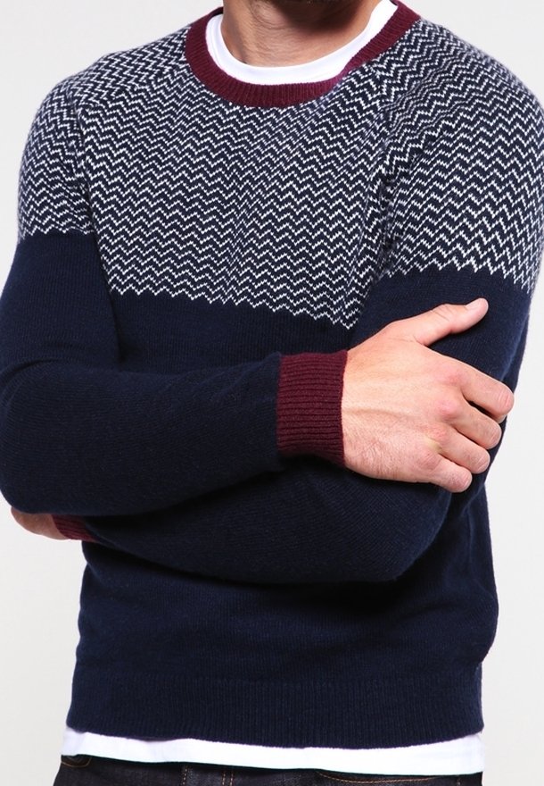 Pull en bleu marine avec un motif en zigzag en blanc et gris en haut. Il possède un col et des poignets côtelés maron. Coupe classique.