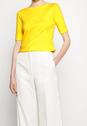 Persona vistiendo una blusa amarilla brillante con cuello bote y mangas hasta el codo, y pantalones blancos de pierna ancha y cintura alta, con una mano en el bolsillo.