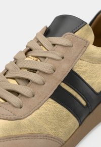 Μπεζ και χρυσό αθλητικό παπούτσι με μαύρη δερμάτινη ρίγα, μπεζ σουέντ λεπτομέρειες και μπεζ υφαντά κορδόνια σε ανοιχτή σόλα, κοντινή λήψη στην περιοχή των κορδονιών.