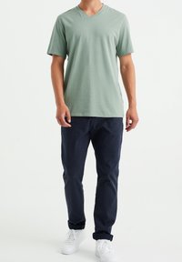 T-shirt vert clair à col en V, manches courtes, porté avec un pantalon bleu marine foncé et des baskets blanches. Tissu lisse, coupe décontractée.
