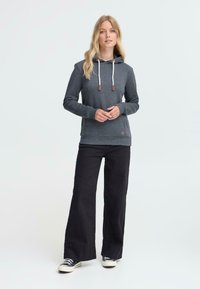 Sweat à capuche gris avec poche kangourou, cordons de serrage, et accents en bois, associé à un pantalon large noir et des baskets noires.