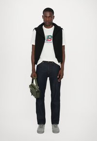 Svart ermeløs genser over en hvit grafisk t-skjorte, mørkeblå jeans, grå joggesko og bærer en grønn crossbody-veske.