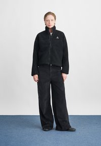 Jachetă din fleece neagră cu fermoar, guler înalt și buzunare frontale; asortată cu pantaloni negri din catifea cei largi și pantofi negri de tip plat. Fundal simplu.