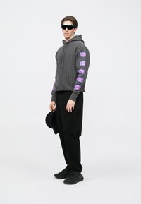 Hanorac gri cu imprimeuri grafice mov pe mâneci, pantaloni negri și adidași, completate de ochelari de soare negri și o șapcă neagră.