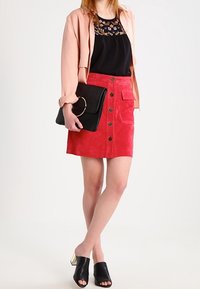Gonna in suede rossa con bottoni frontali e tasche, abbinata a una blusa nera con ricami, una giacca rosa chiaro e una pochette nera. Mule nere con punta aperta.