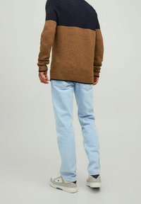 Hombre vestido con un suéter de punto marrón y azul oscuro, jeans azul claro y zapatillas grises, de pie de espaldas frente a un fondo liso.