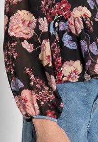 Blusa floral em tecido preto transparente, com flores vermelhas, rosas e azuis. Mangas bufantes com punhos franzidos e um design de cintura peplum.