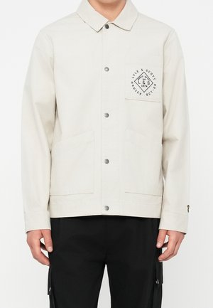 Personne portant une veste beige clair boutonnée avec trois poches devant et un logo noir imprimé sur la poche poitrine gauche, associée à un pantalon noir.