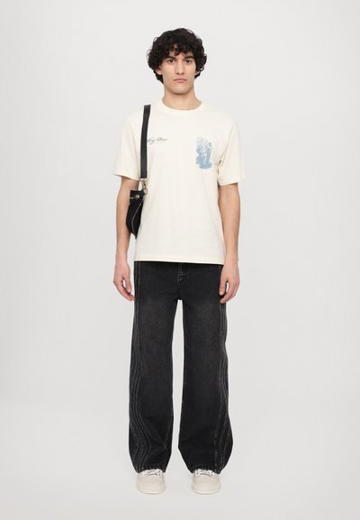 T-shirt en coton crème avec impression graphique bleue, associé à un jean noir ample avec des coutures latérales contrastées et des chaussures blanches.